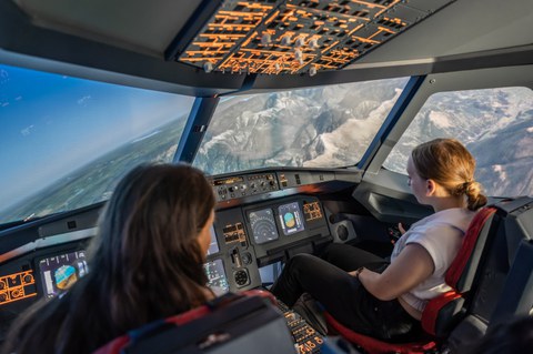 Zwei Schülerinnen sitzen im Cockpit eines Flugsimulators und steuern das Flugzeug. Das Flugzeug befindet sich in Schräglage und überfliegt Gebirge.