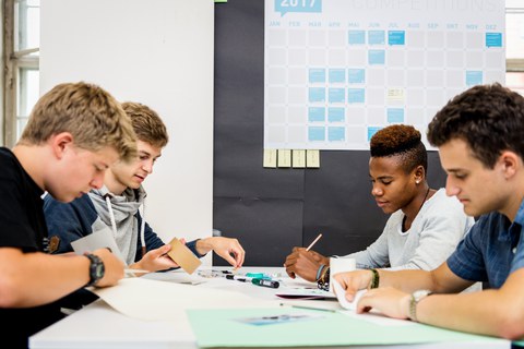 Zu sehen ist ein Foto von vier Jungen an einem Tisch. Sie halten Klebeband, eine Schere, einen Stift und Papier in den Händen. Auf dem Tisch befinden sich weitere Büroutensilien. Im Hintergrund ist ein Wandkalender zu erkennen.