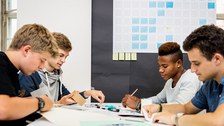 Zu sehen ist ein Foto von vier Jungen an einem Tisch. Sie halten Klebeband, eine Schere, einen Stift und Papier in den Händen. Auf dem Tisch befinden sich weitere Büroutensilien. Im Hintergrund ist ein Wandkalender zu erkennen.