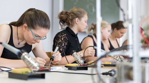 The picture shows a photo of teenage girls soldering in a laboratory.