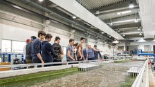 Zu sehen ist ein Foto. Eine Jungengruppe steht in einer großen Halle hinter dem Modell eines Flusses. Die Blicke sind auf das Modell gerichtet. Eine Frau zeigt auf dieses Modell und spricht.