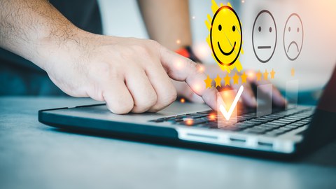 Eine Hand, die auf einer Laptoptastatur drückt. Es erscheinen Smileys, wovon der glückliche farblich hervorgehoben ist. 