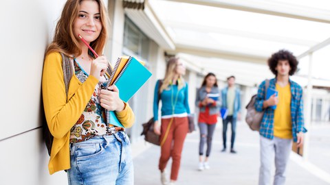 Foto einer jungen Frau, die Stift und Hefte in der Hand trägt und an einer Wand lehnt; verschwommen im Hintergrund: vier junge Frauen und Männer