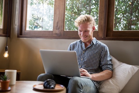 Ein Mann, der auf einem Sofa sitzt mit einem Laptop auf dem Schoß und lächelt.