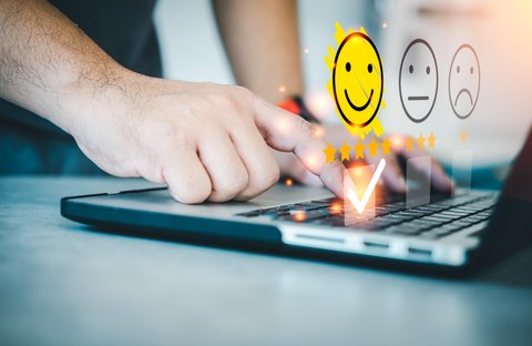 Eine Hand, die auf einer Laptoptastatur drückt. Es erscheinen Smileys, wovon der glückliche farblich hervorgehoben ist. 