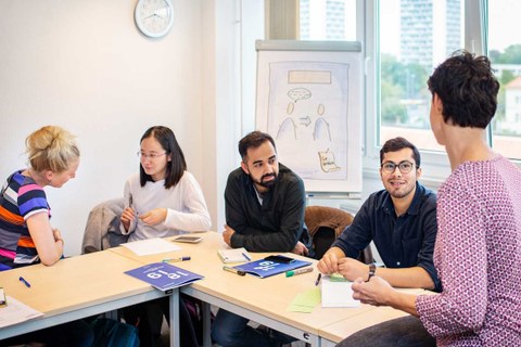 Das Foto zeigt vier Studierende die an einem Tisch sitzen. Im Vordergrund ist eine Referentin zu sehen. Im Hintergrund steht ein Wideboard.