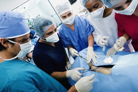 Das Foto zeigt fünf Personen in einem Operationssaal. Sie tragen Haarnetze, Handschuhe und Mundschutz. Sie stehen um einen Patienten herum und sin bereit, einen Eingriff vorzunehmen.
