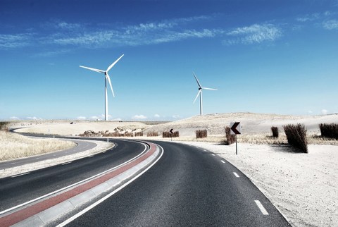 Das Foto zeigt eine Straße, die sich durch eine Wüstenlandschaft schlängelt. Im Hintergrund erkennt man zwei Windräder. 
