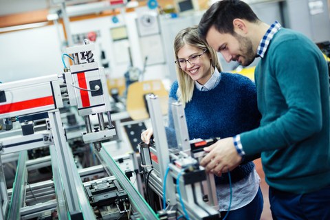 Das Foto zeigt zwei Studierende, die an einer hochtechnisierten Maschine stehen. Die Studierenden lächeln. 