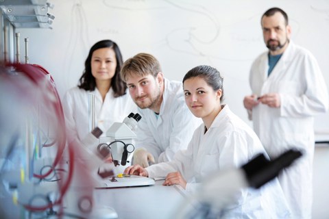 Das Foto zeigt vier Personen in Laborkitteln an einem Tisch. Auf dem Tisch steht ein Mikroskop. Die Personen schauen alle in die Kamera. 