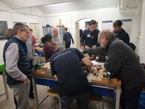 You can see several people in a room watching one person repair an old turntable.