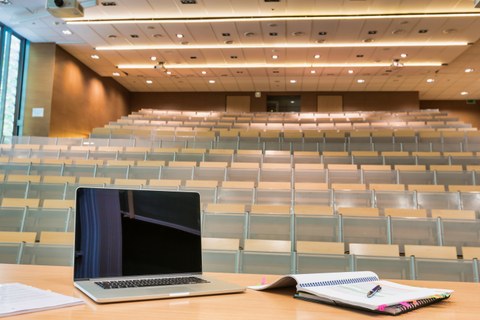 Photo. Lecture hall. Foreground: Surface of a lecturer's desk, on the left a notepad and an opened laptop with a black screen, on the right an opened appointment calendar with colorful sticky notes on the side. A ballpoint pen lies on top. Background: Empty lecture hall (slanted movie theater architecture) with lighting from the ceiling. Lecturer's table and chairs in light wood color; audience tables in light gray.