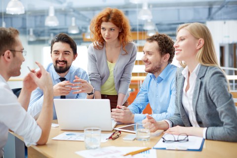 Foto: Eine Gruppe von Mitarbeitenden sitzen an einem Tisch, in der Mitte steht ein Laptop. Sie diskutieren miteinander.