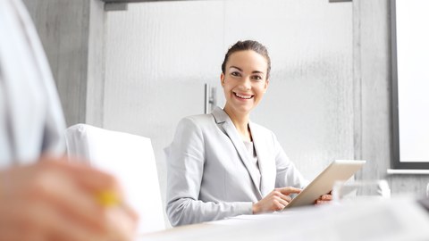 Foto einer lächelnden Business-Frau. Sie sitzt am Schreibtisch mit einem Tablet in der Hand und lächelt freundlich. 