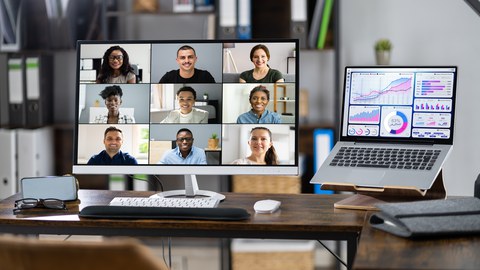 Foto: Ein großer Monitor steht auf einem Schreibtisch, darin zu sehen ist eine virtuelle Online-Videokonferenz auf mehreren Teilnehmenden. Daneben ein Laptop mit Diagrammen auf dem Display. 