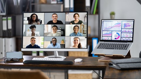 Foto: Ein großer Monitor steht auf einem Schreibtisch, darin zu sehen ist eine virtuelle Online-Videokonferenz auf mehreren Teilnehmenden. Daneben ein Laptop mit Diagrammen auf dem Display. 