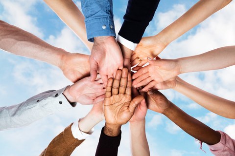 Photo of many outstretched hands of different skin colors lying crossed over each other, in the background is the sky