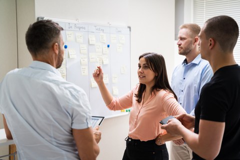 Foto: Ein Projektteam steht vor einem Kanban White Board und diskutiert den Aufgabenplan.