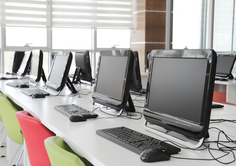 Foto eines Computerraums ohne Menschen, in einer Reihe stehen Monitore und Tastaturen. Es gibt grüne und rote Stühle. 