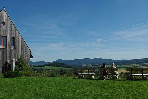 Foto. Im Vordergrund eine grüne Wiese und Bänke, auf denen Menschen sitzen. Im Hintergrund eine hügelige Landschaft und blauer Himmel. Die Menschen betrachten die Landschaft in der Ferne.