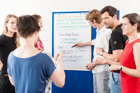 6 people at a workshop are gathered round a pinboard. A man is pointing at the board. The others are facing a woman who has her back to the camera.