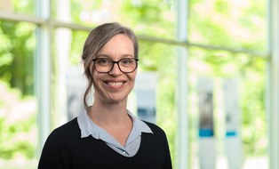 Portraitfoto von Beatrice Schlegel. Sie trägt lange dunkle Haare zusammengesteckt und eine Brille, einen dunklen Pullover, aus dem ein hellblauer Hemdkragen herausschaut.