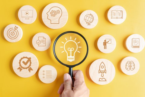 Foto einer Glühbirne, die unter einer Lupe betrachtet wird. Um die Glühbirne herum liegen rund Holzplättchen mit verschiedenen Icons wie ein Globus, eine Rakete oder ein Buch.