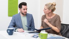 A woman and a man are sitting at a desk, looking at a folder. In front of them are two TUD coffee cups.