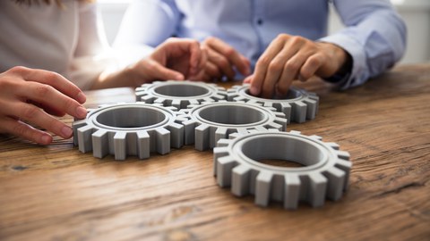oto von Zahnrädern, die auf einem Tisch liegen und ineinander greifen. Um den Tisch sitzen mehrere Personen, die ihre Hände an den Zahnrädern haben.