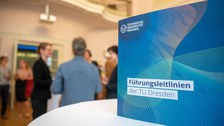 Die Broschüre Führungsleitlinien der TU Dresden steht auf einem Tisch, im Hintergrund sind Menschen verschwommen zu erkennen.