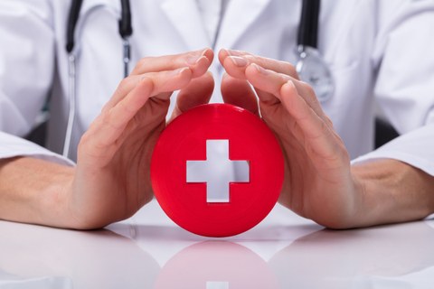 Auf dem Foto ist ein rundes rotes Symbol zu sehen, welches ein Kreuz in der Mitte hat. Hinter dem Symbol sitzt ein Arzt mit einem Arztkittel und umrahmt das Symbol mit seinen Händen.