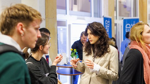Foto: Eine Person in hellem Blazer spricht gestikulierend mit jungen Menschen auf einer Messe. 