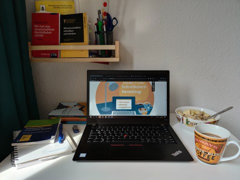A laptop stands open on a desk, with a graphic on it that reads "2.09. 7-12 a.m.: Shrine Start Morning." In the background, books for scientific reading and writing.