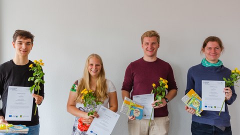 Robin Wratil, Marina Hüttich, Johannes Reimer und Jochen Diepelt schauen lachend in die Kamera. Sie halten in ihren Händen eine gelbe Blume, eine Urkunde und ein Geschenk. Von links nach rechts stehen nebeneinander:  