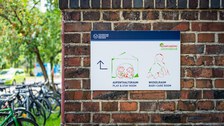 Foto einer Backstein mit einem Schild. Das Schild ist ein Wegweiser des Campusbüro „Uni mit Kind“ zum Aufenthaltsraum und zum Wickelraum