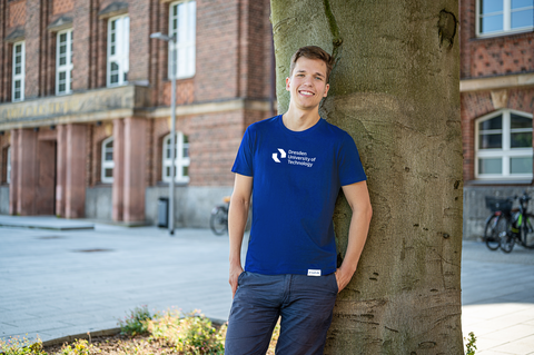 A young man is standing on the TUD campus leaning against a tree. He is wearing a blue TUD shirt and jeans.