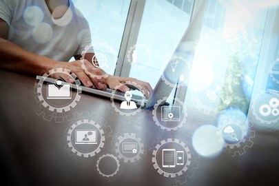 Photo of a person working on a laptop. In the foreground are different sized gears with symbols in the center such as a cell phone, tablet, cloud and magnifying glass. 