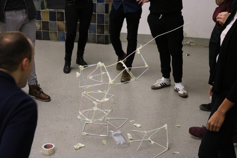 Foto vom Teambuilding. Eine Gruppe Studierender steht im Kreis um eine abstrakte Figur gebastelt aus Strohhalmen herum.