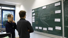 Foto von zwei Studenten, die vor einer beschrifteten Tafel stehen und nachdenken.