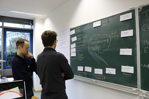 Foto von zwei Studenten, die vor einer beschrifteten Tafel stehen und nachdenken.