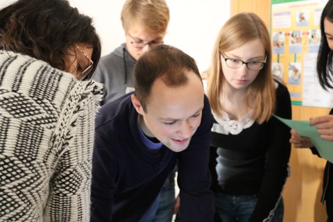 Foto der Teamarbeit. Studierende stehen beisammen und schauen auf etwas hinab, das sich außerhalb des Fotos befindet.