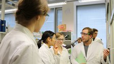 Foto von der Chemie Laborzeit. Studierende stehen mit Arbeitskitteln und Schutzbrillen in einem Labor und unterhalten sich.