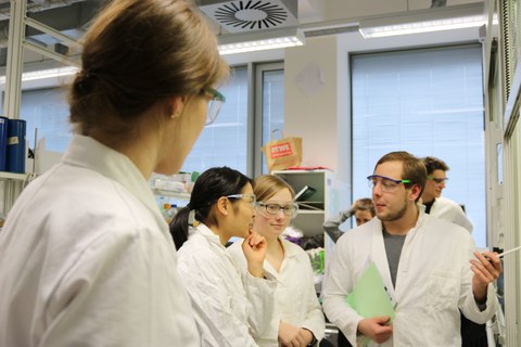 Foto von der Chemie Laborzeit. Studierende stehen mit Arbeitskitteln und Schutzbrillen in einem Labor und unterhalten sich.