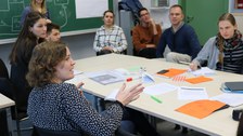 Foto von der Profirunde. Studierende sitzen um einen Tisch auf dem Arbeitsmaterialien wie Blätter und Stifte liegen. Eine Studentin spricht mit jemandem, der sich außerhalb des Fotos befindet.
