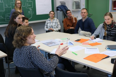 Foto von der Profirunde. Studierende sitzen um einen Tisch auf dem Arbeitsmaterialien wie Blätter und Stifte liegen. Eine Studentin spricht mit jemandem, der sich außerhalb des Fotos befindet.