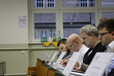 Foto von der Abschlusspräsentation. Die Jury sitzt in einer Reihe in einem Hörsaal und füllt Bewertungsbögen aus.