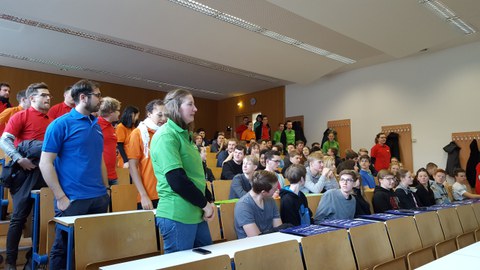 Foto von Studierenden in einem Hörsaal. Die Team- und Fachcoaches stehen und tragen T-Shirts in ihren Teamfarben. Ihre jeweiligen Gruppen sitzen neben ihnen in den Stuhlreihen.