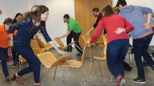 Foto von Studierenden während einer Teambuilding Methode. Sie stehen neben einem Stuhlkreis und halten jeweils einen Stuhl mit einer Hand fest.