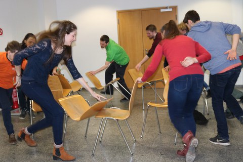 Foto von Studierenden während einer Teambuilding Methode. Sie stehen neben einem Stuhlkreis und halten jeweils einen Stuhl mit einer Hand fest.