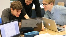 Foto von drei Studenten. Sie sitzen um einen Tisch herum und arbeiten an ihren Laptops. Auf dem Tisch liegen weitere Arbeitsmaterialien wie Stifte und Blätter.